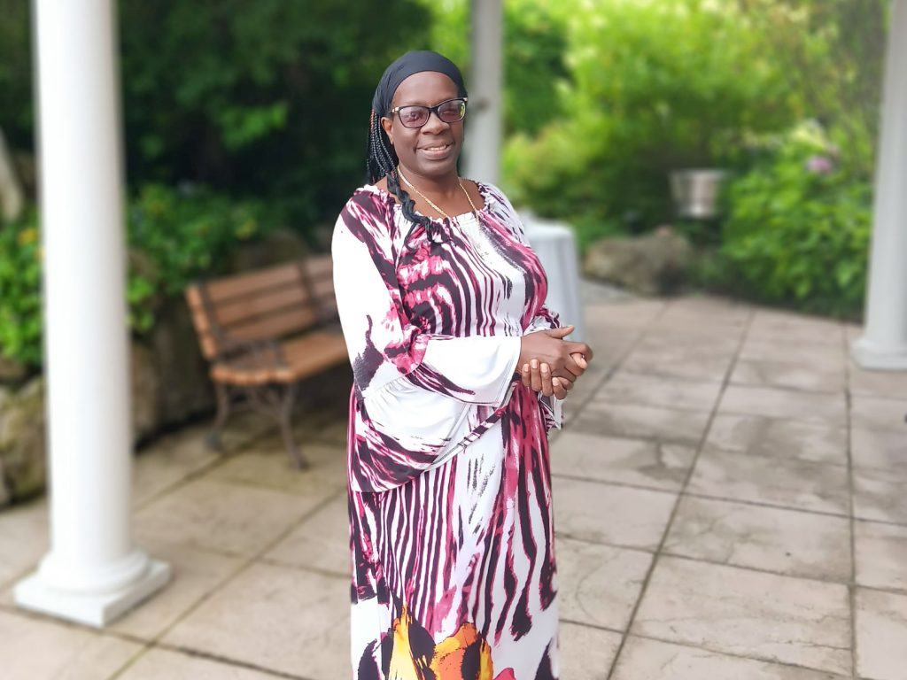 Photo of a smiling woman wearing glasses and a white and pink dress, standing in a park. 