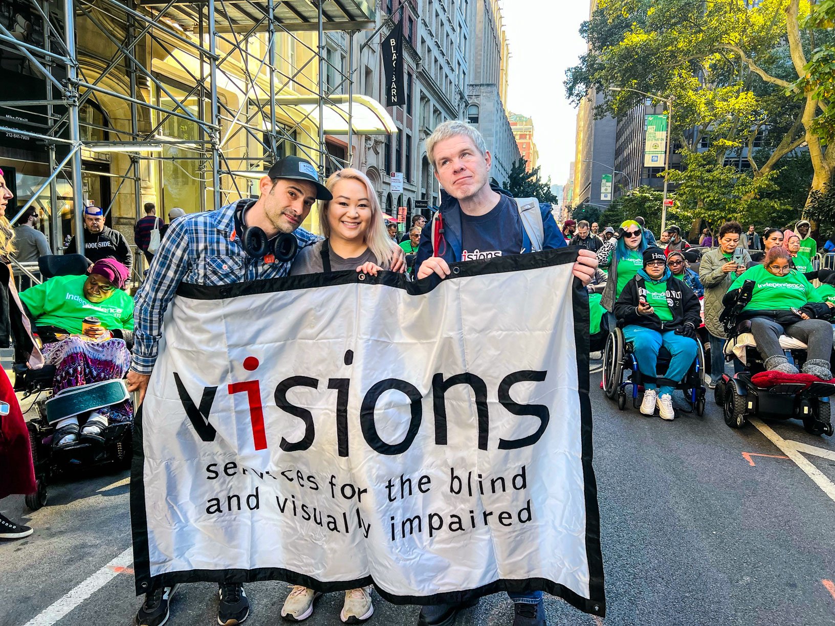 Photo of three VISIONS staff members holding a large VISIONS banner as they walk along an outdoor parade.