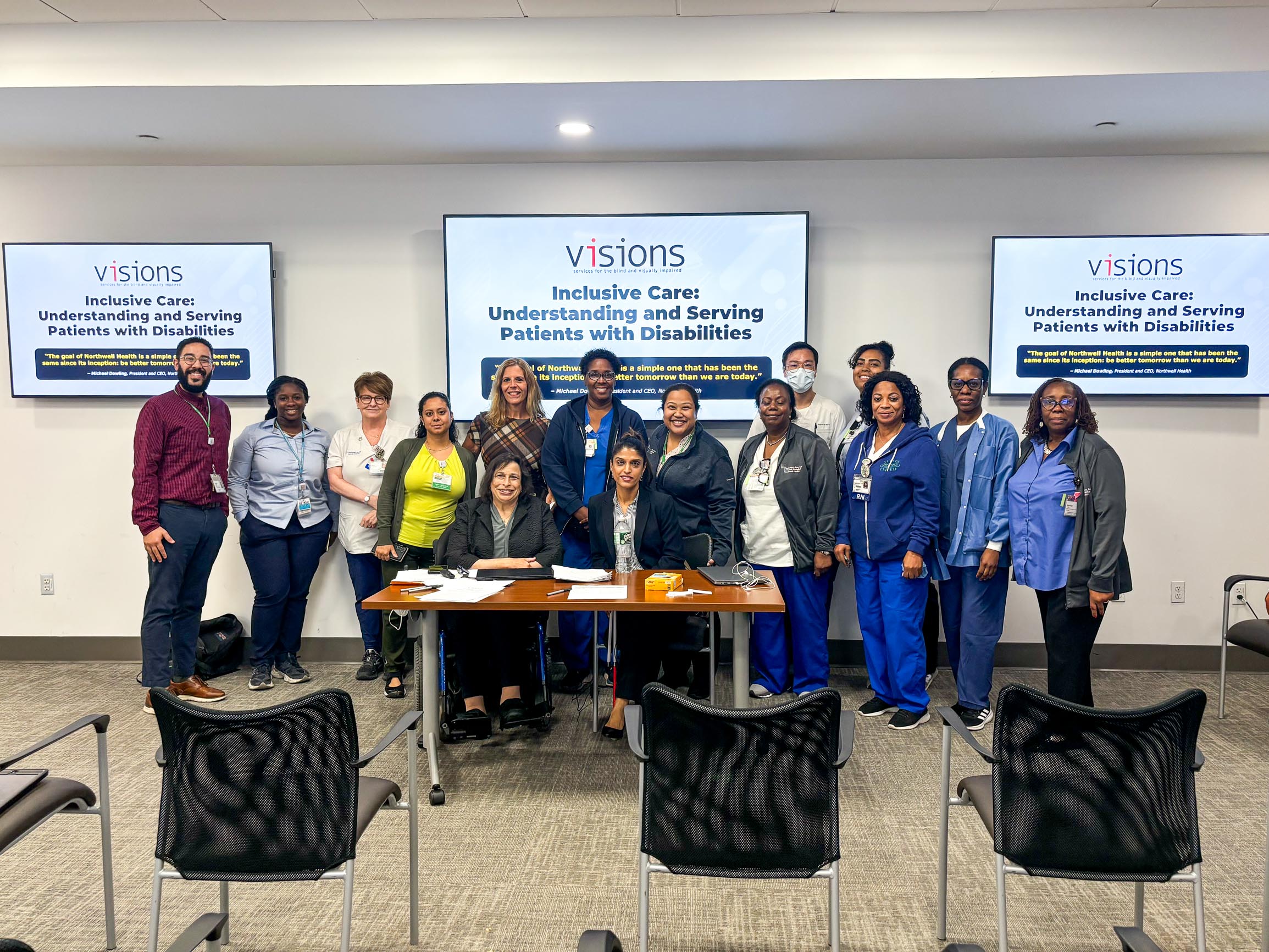 Photo of a group of VISIONS and medical staff standing together underneath a VISIONS Equal Access to Healthcare PowerPoint projected on several TV screens.